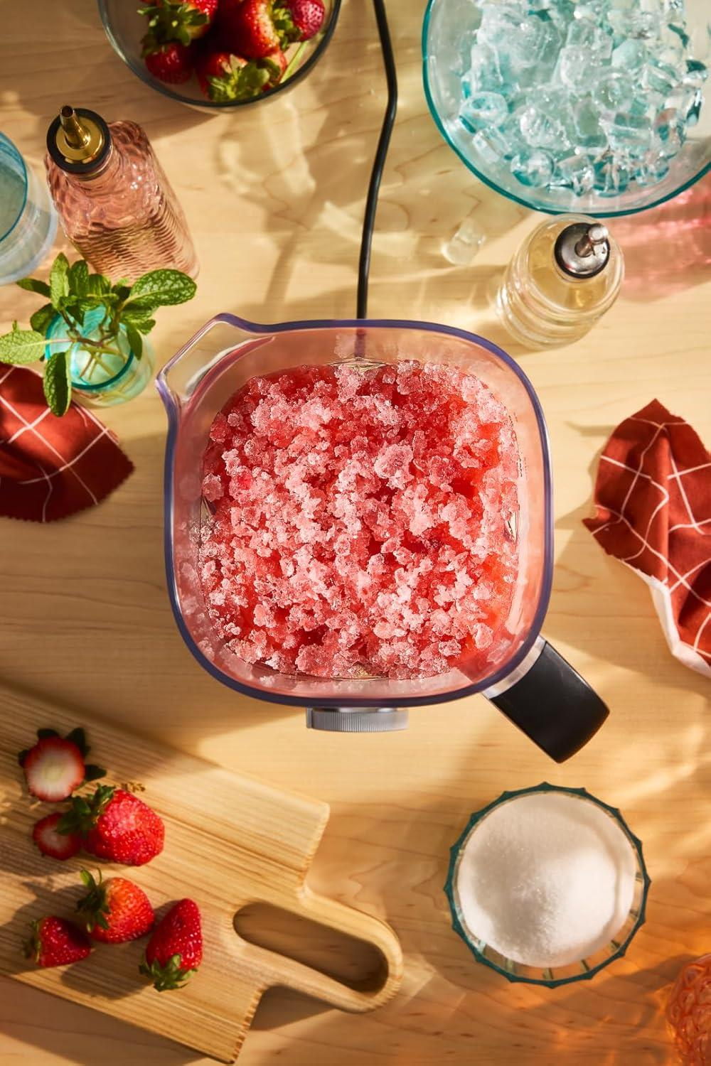 Ein Mixerbehälter mit zerstoßenem Eis und Erdbeeren, umgeben von Zutaten.