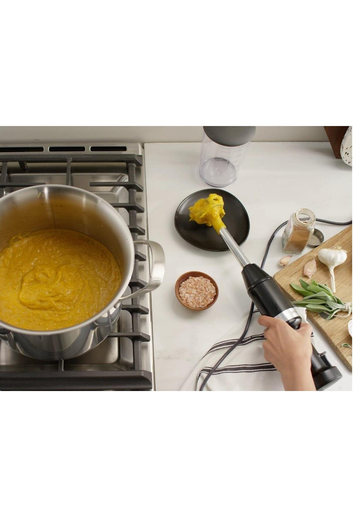 Een hand houdt een zwarte staafmixer boven een kom met gele puree. Daarnaast een pan met puree op het fornuis.