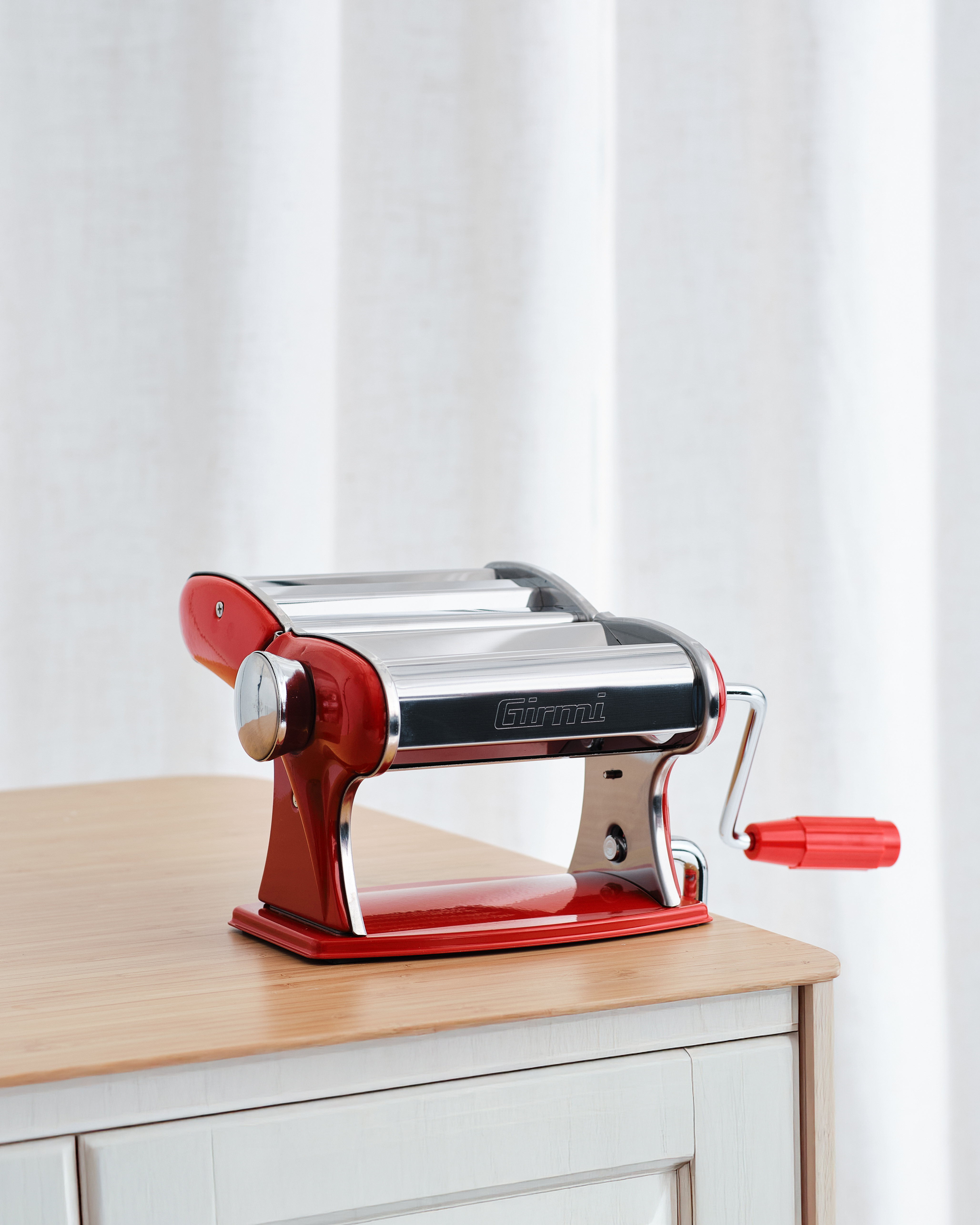 Rote Nudelmaschine mit silbernen Walzen und Kurbel auf einem Holztisch.