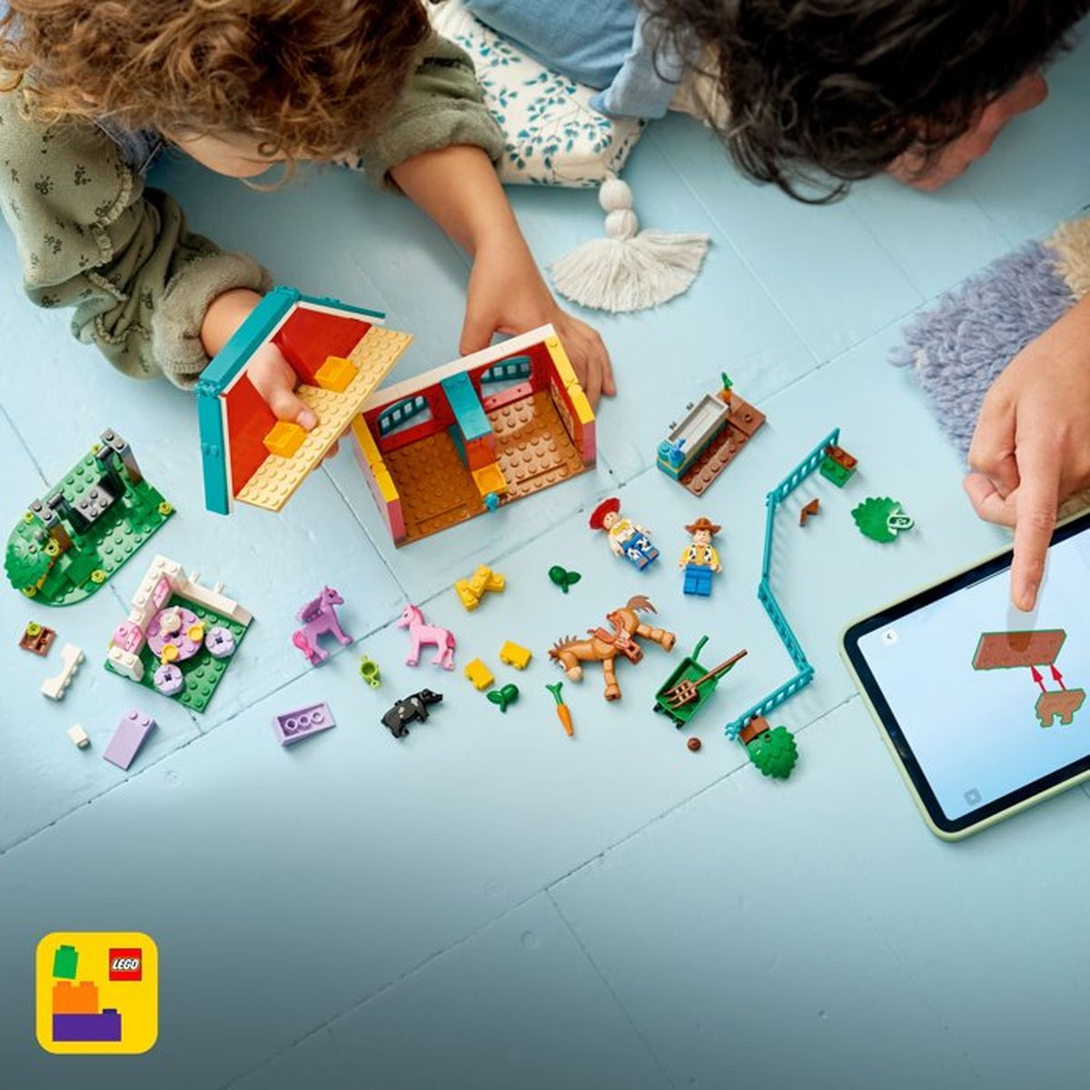 Dzieci budują z klocków Lego, w tym konie, figurki i dom. Tablet pokazuje instrukcje.