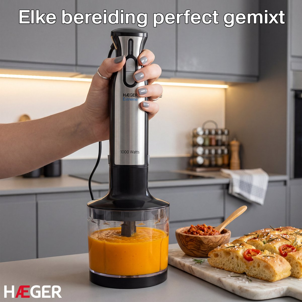 Een hand houdt een zilveren staafmixer met zwarte handgreep boven een bak met oranje vloeistof.