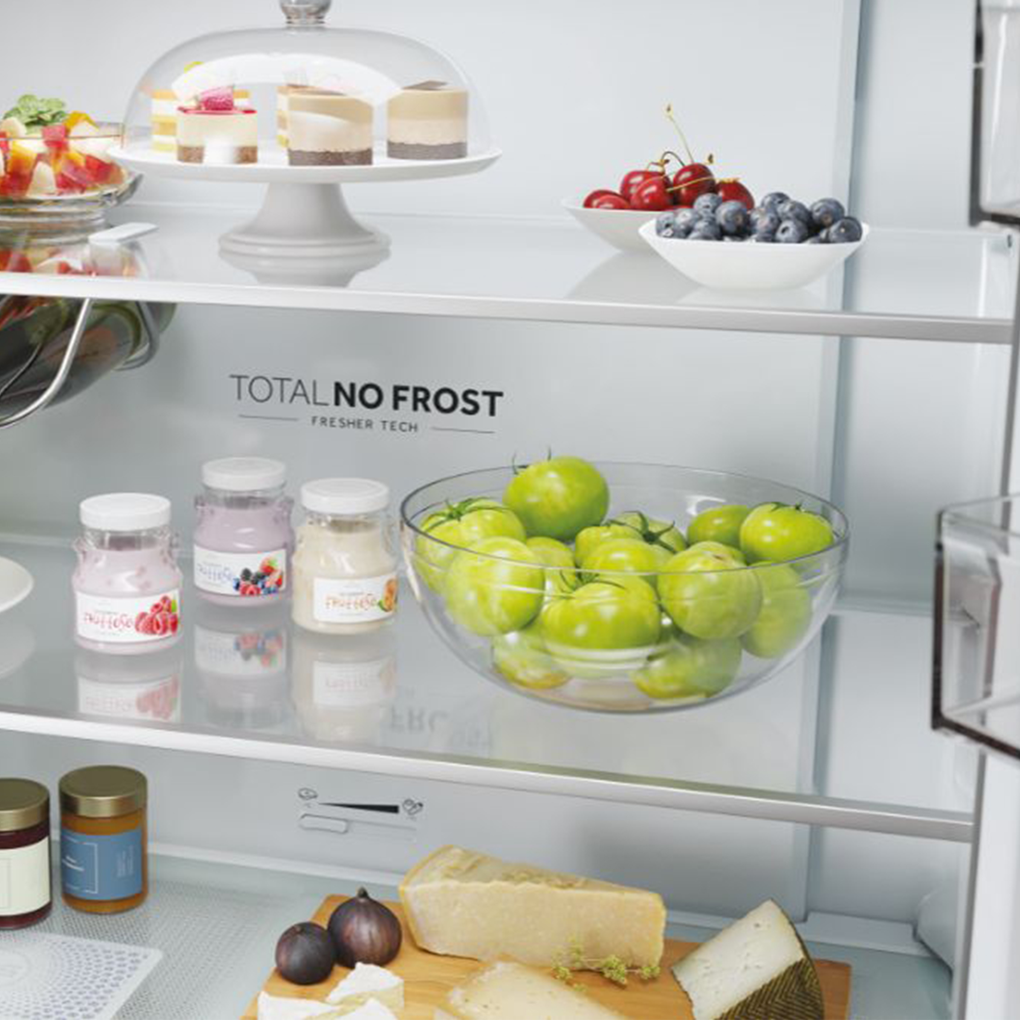 Una repisa de refrigerador con frutas, pasteles, yogur, tomates y queso.