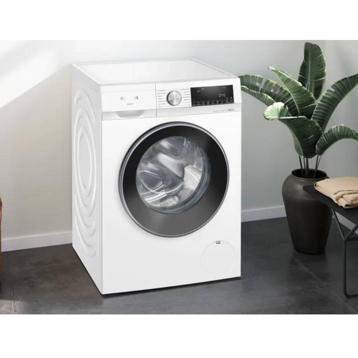 Lavadora blanca con puerta negra y pantalla digital, junto a una planta.