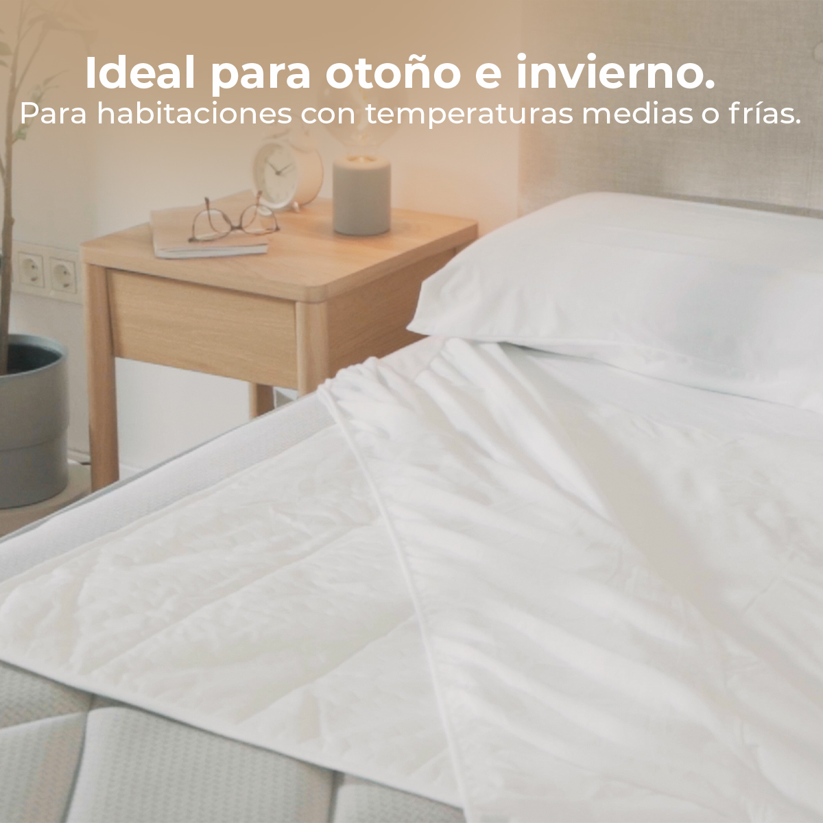 Un comodino con sveglia, candela e occhiali accanto a un letto bianco.