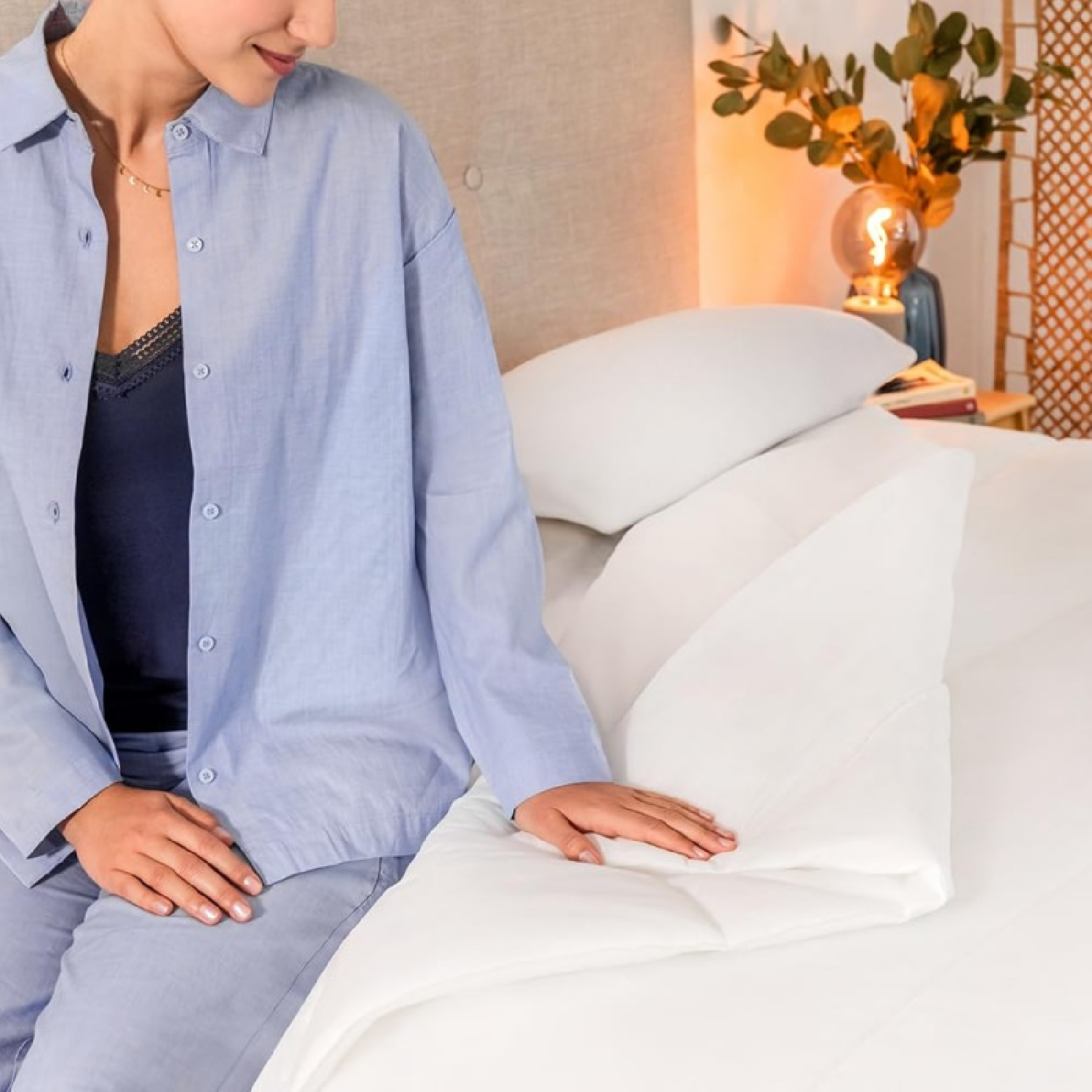 Donna in pigiama blu seduta su letto, tocca piumone bianco piegato. Lampada comodino accesa.