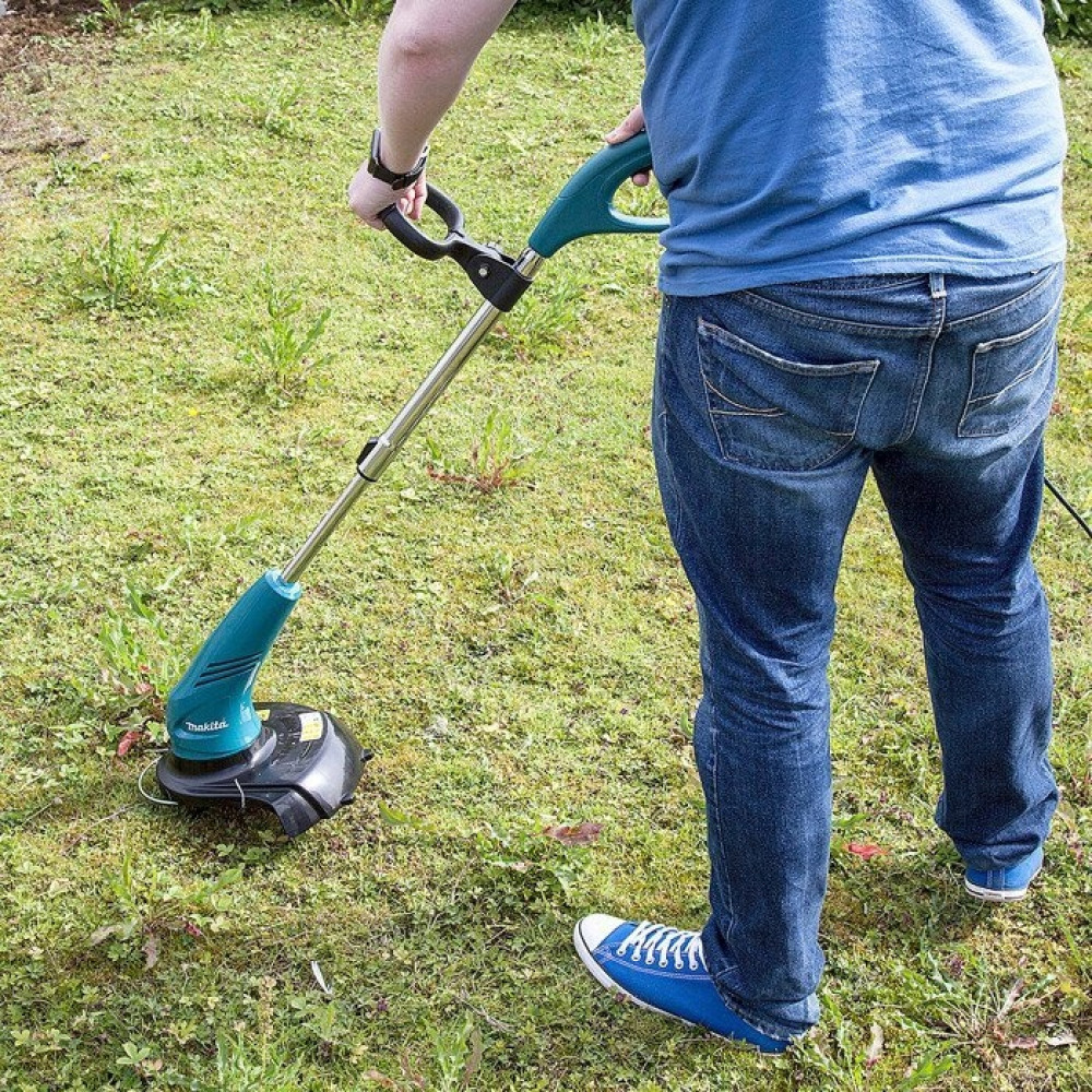 Osoba w dżinsach kosi trawę podkaszarką w ogrodzie.