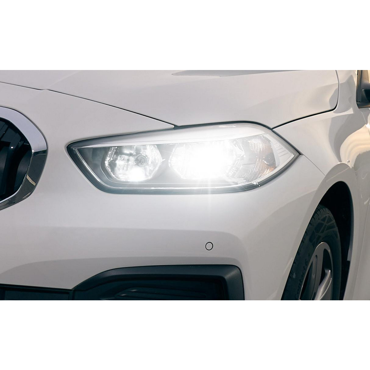 Vorderansicht eines weißen Autos mit eingeschalteten Scheinwerfern und Chromgrill.