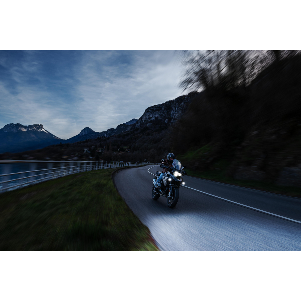 Motorradfahrer auf kurviger Straße im Dämmerlicht, Berge und See im Hintergrund.