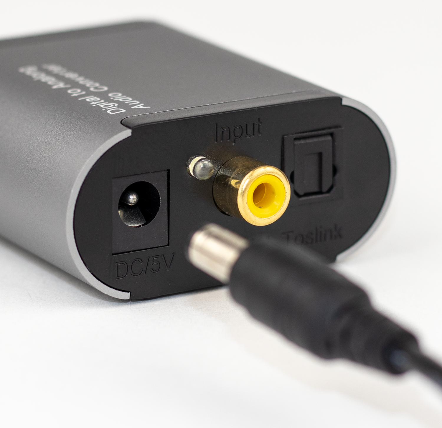 Convertidor de audio gris con entrada DC/5V, entrada RCA y puerto Toslink. Cable negro enchufado.