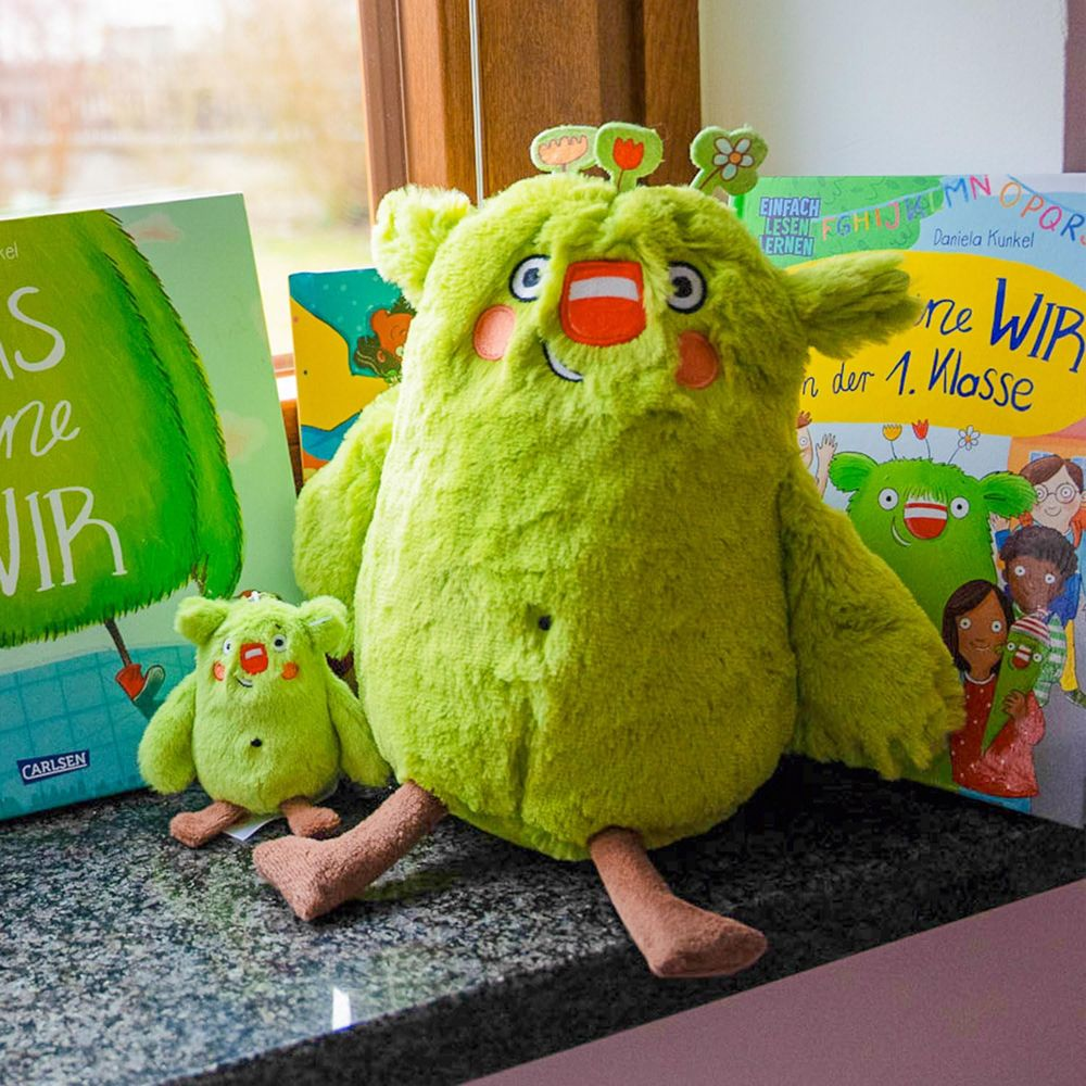 Deux monstres en peluche verts sont assis à côté de deux livres pour enfants sur une surface sombre.