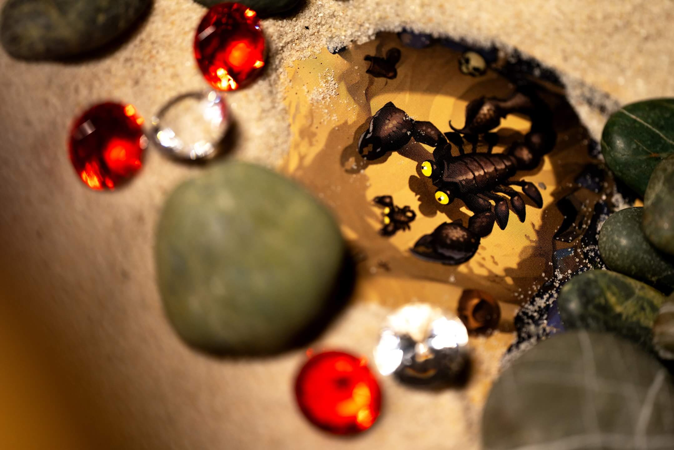 Un scorpion jouet aux yeux jaunes sur un tissu à motifs, entouré de sable et de pierres.
