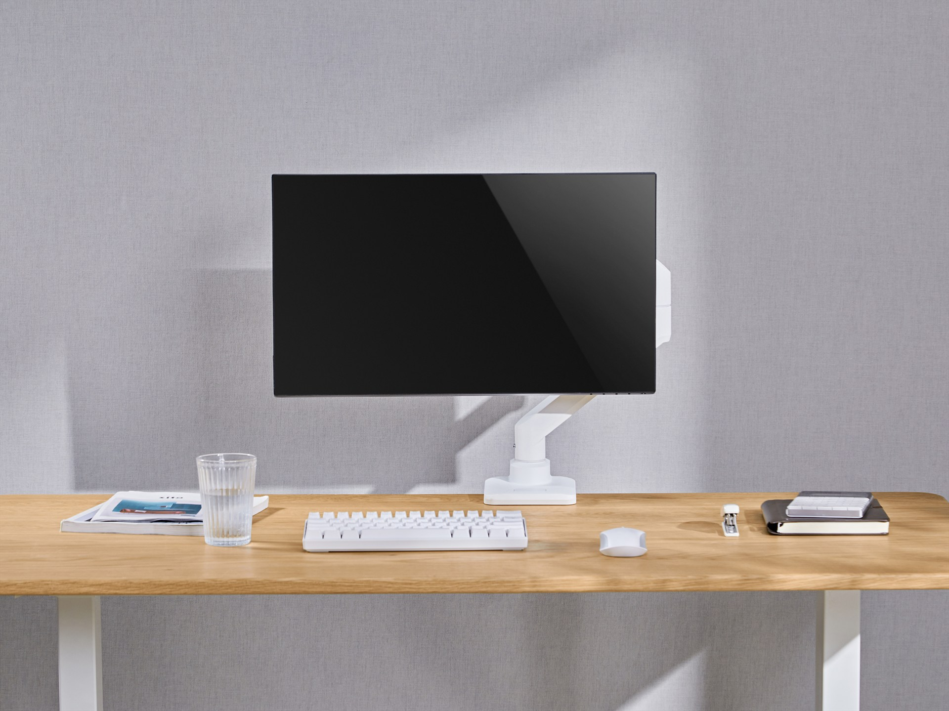 Ein weißer Computermonitor auf einem verstellbaren Arm über einem Holztisch mit Tastatur, Maus und Glas.