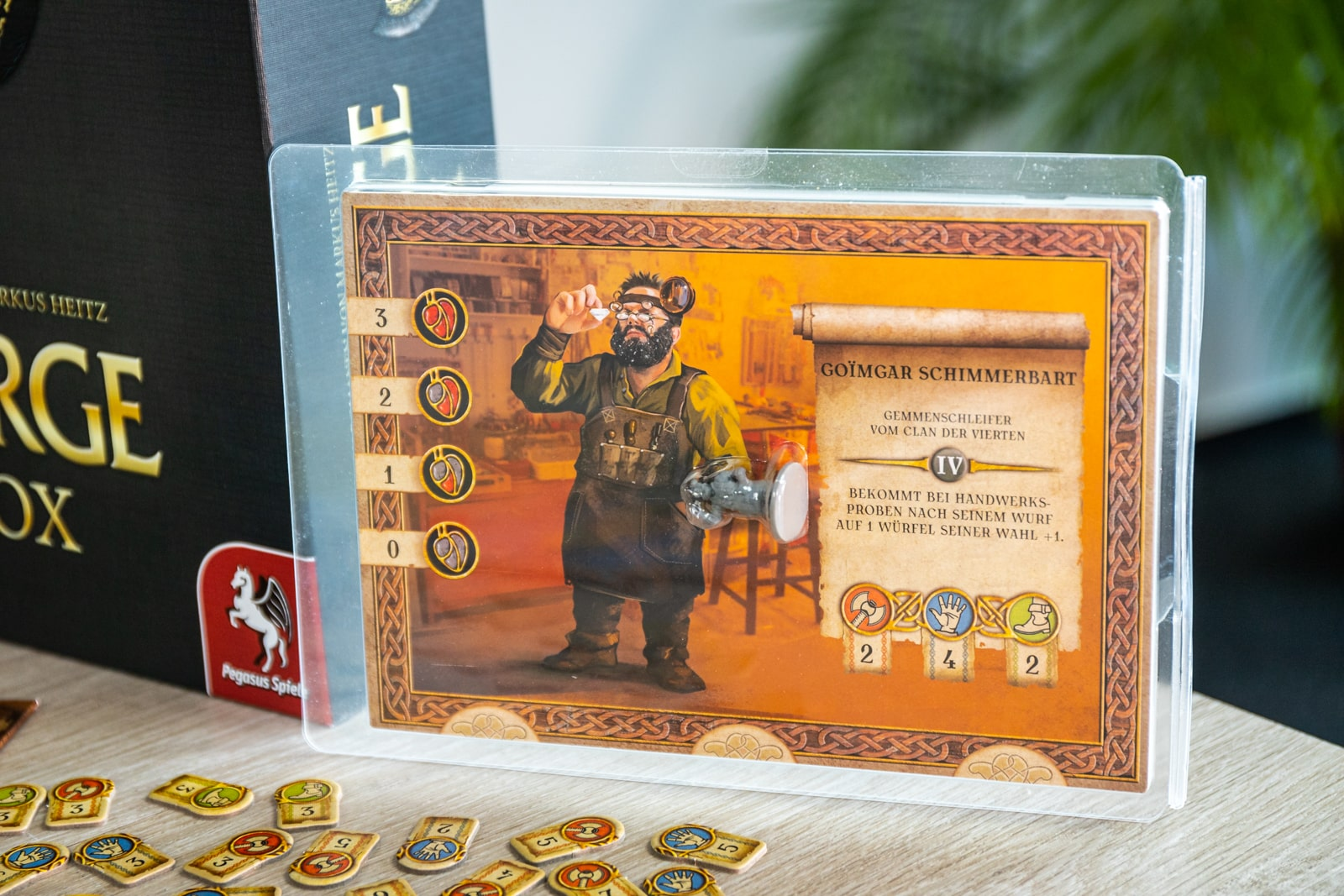 Eine Spielkarte zeigt einen bärtigen Zwergenhandwerker mit Werkzeugen und Werten. Eine Spielbox ist sichtbar.
