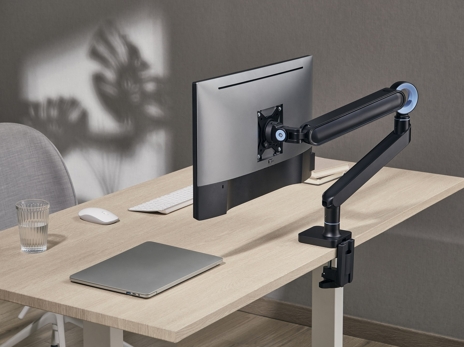 Ein Monitorarm hält einen Bildschirm über einem Schreibtisch mit Laptop, Maus und Glas Wasser.