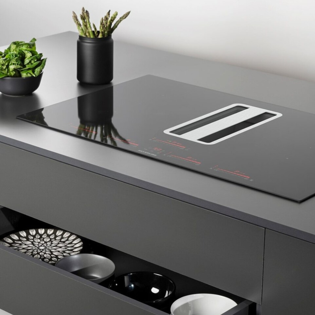 Piano cottura a induzione nero con cappa integrata. Accanto, ciotola con insalata e asparagi.
