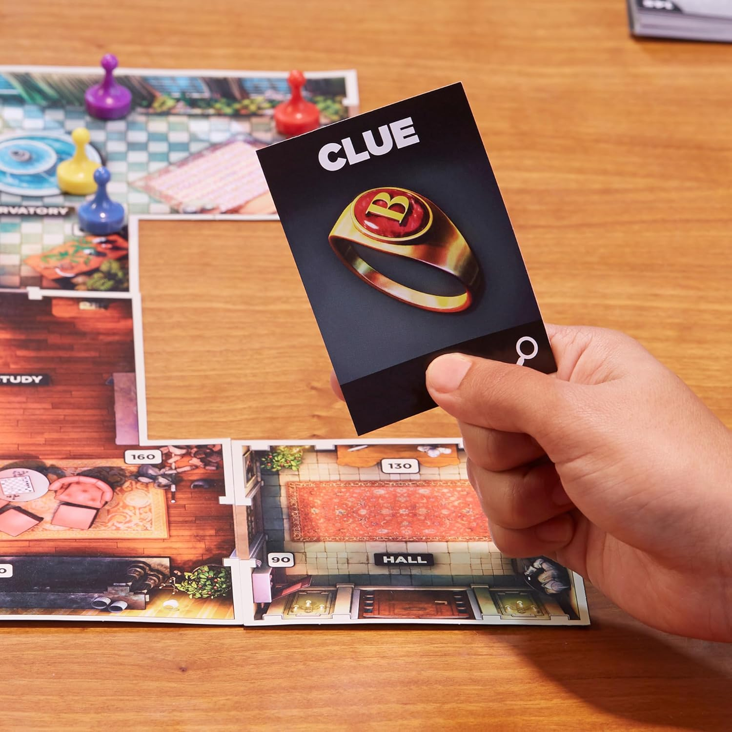 Eine Hand hält eine CLUE-Karte mit einem Ring auf einem Spielbrett mit Spielfiguren.