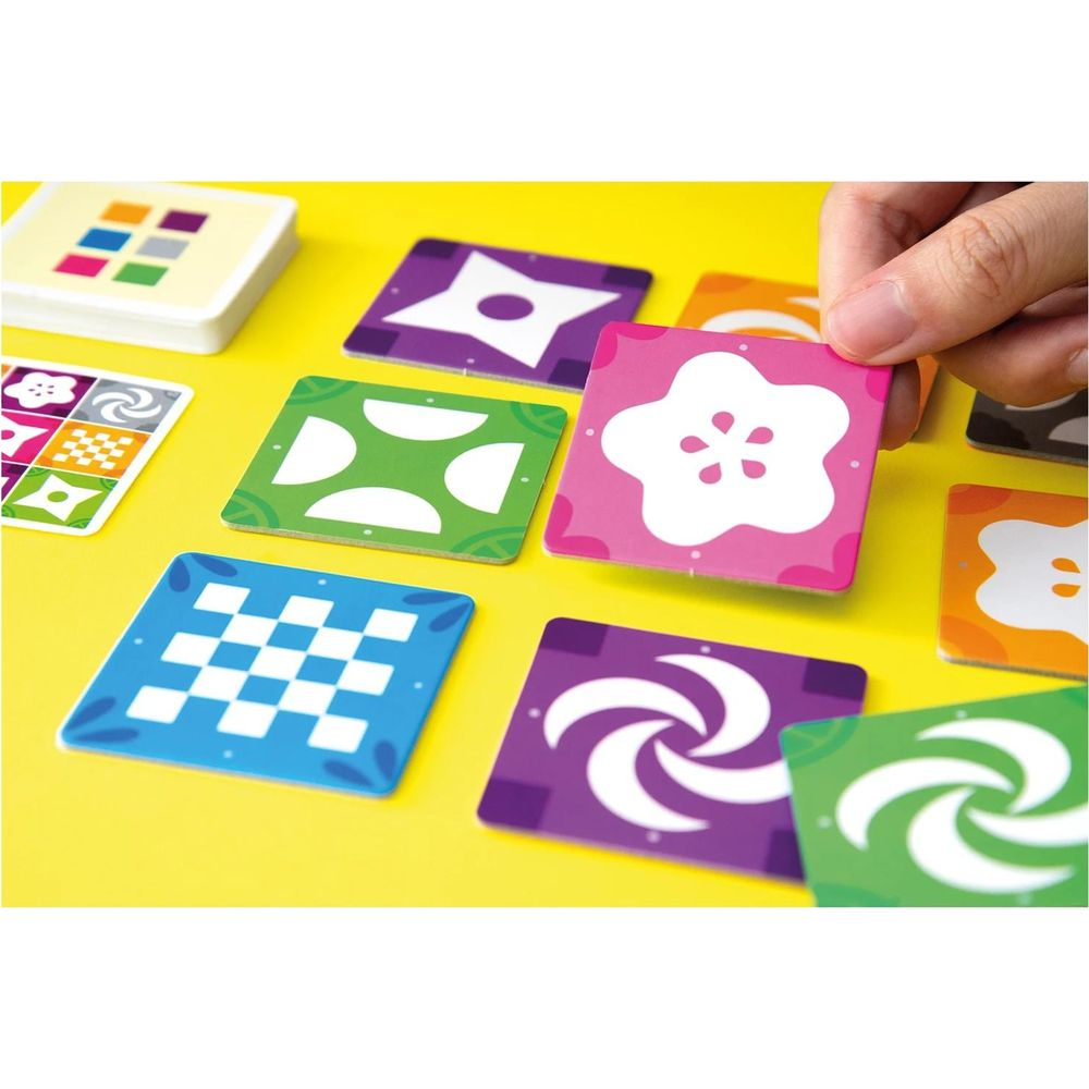 Bunte Spielkarten mit Mustern auf gelbem Hintergrund. Eine Hand hält eine pinke Karte.