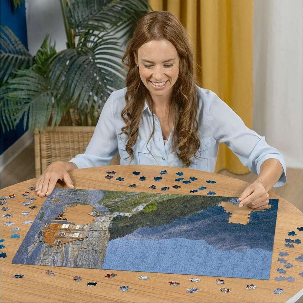 Femme assemblant un puzzle d'une maison sur une falaise surplombant l'eau bleue.