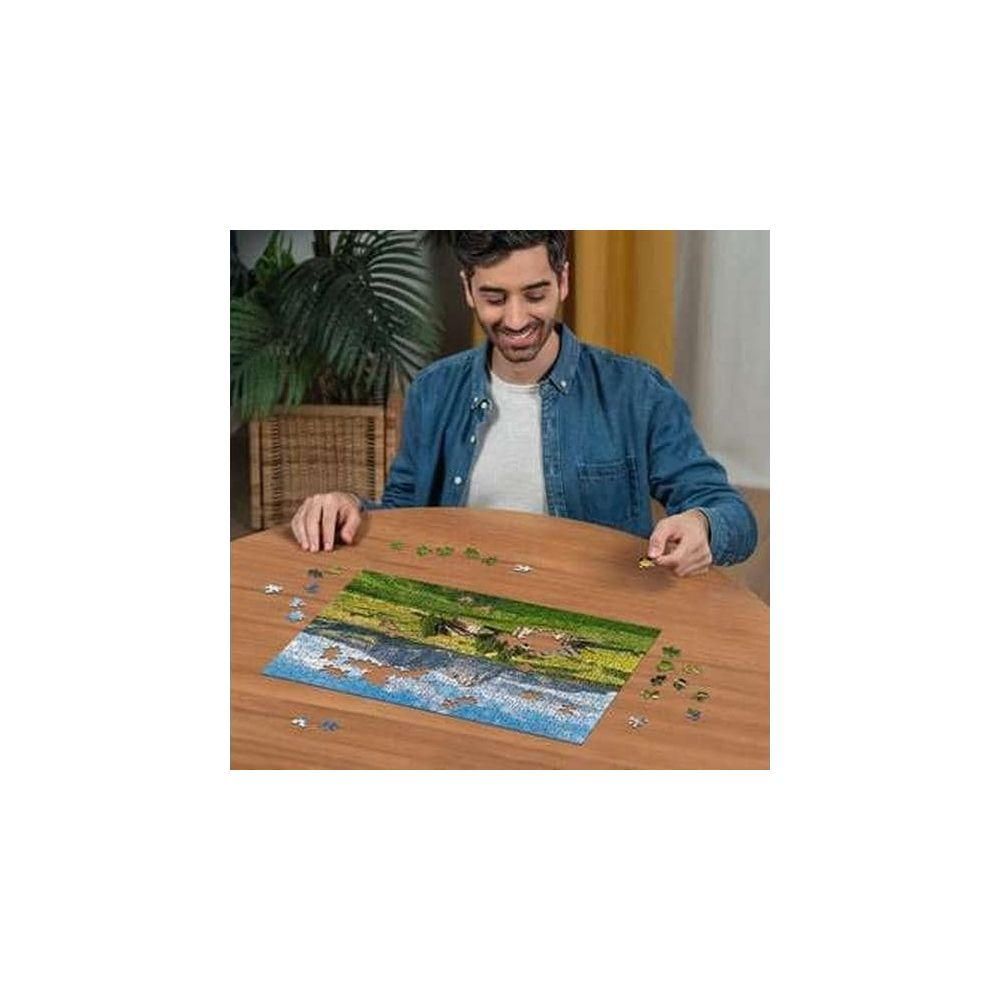 Uomo in camicia di jeans monta un puzzle paesaggistico su tavolo di legno.