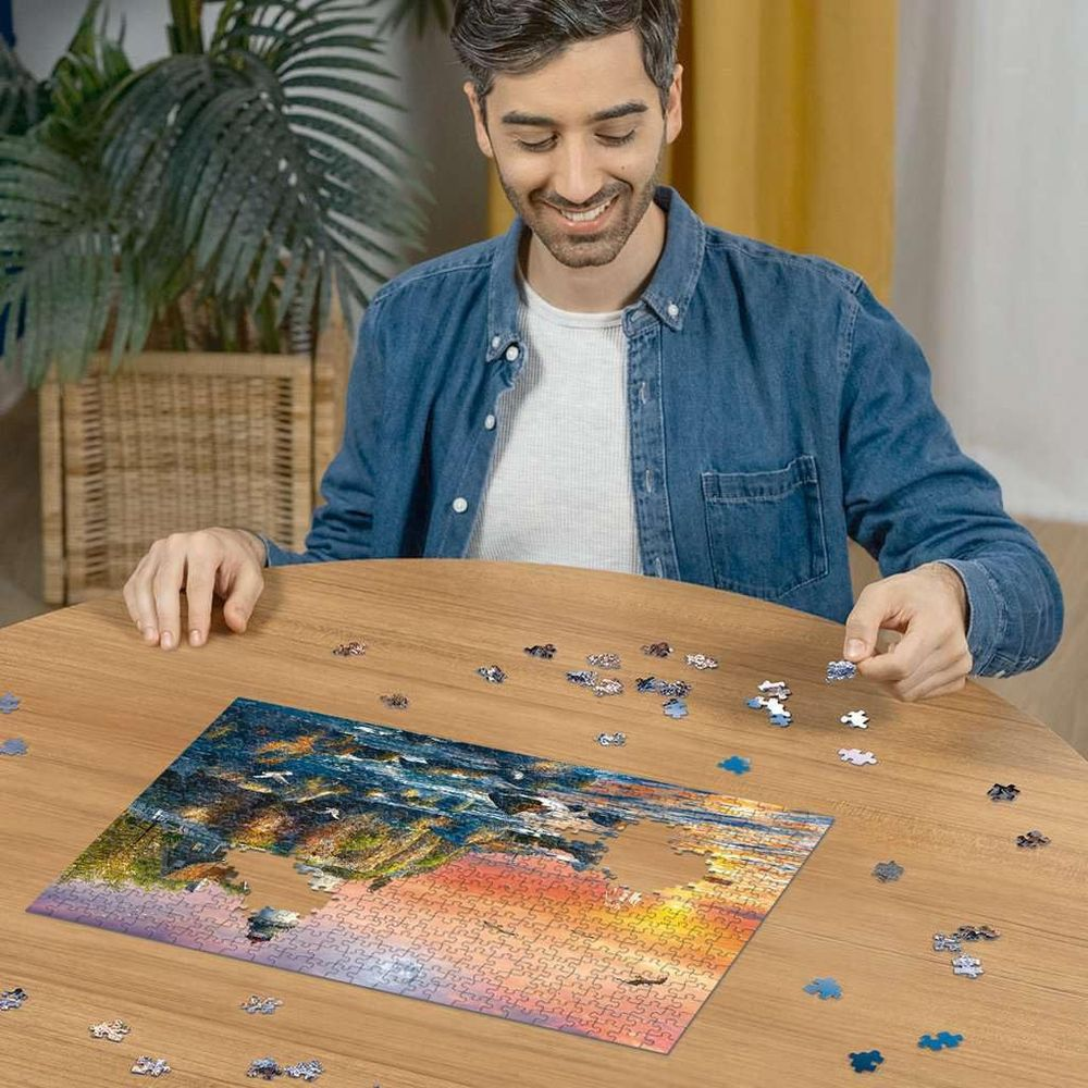 Uomo che assembla un puzzle di un tramonto sull'acqua con pezzi sparsi.