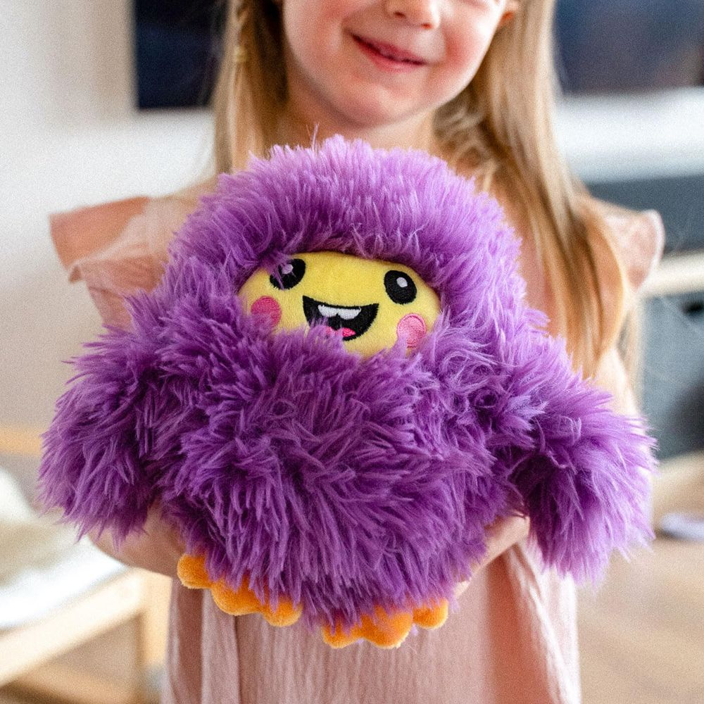 Une fille tient un monstre violet et pelucheux avec un visage jaune et des pieds oranges.
