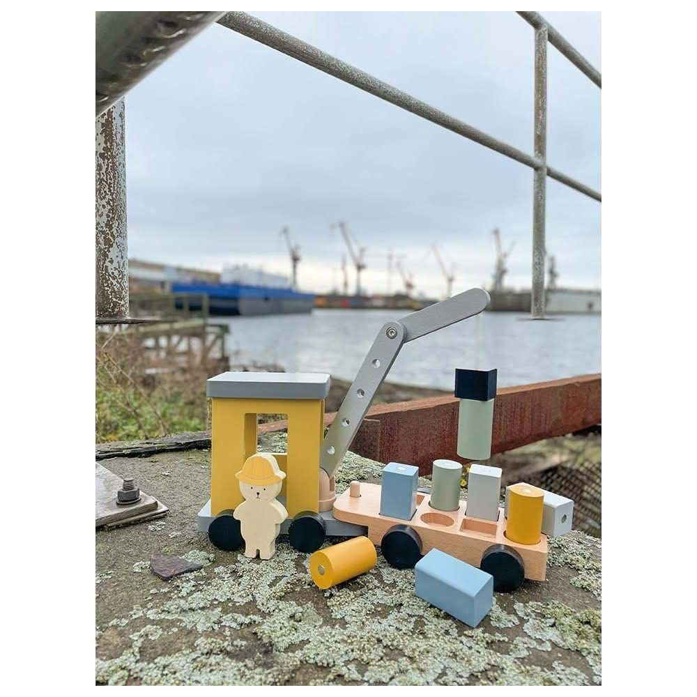 Hölzerne Spielzeugkran mit Blöcken und Figur auf einem Dock, mit Schiffen und Kränen im Hintergrund.
