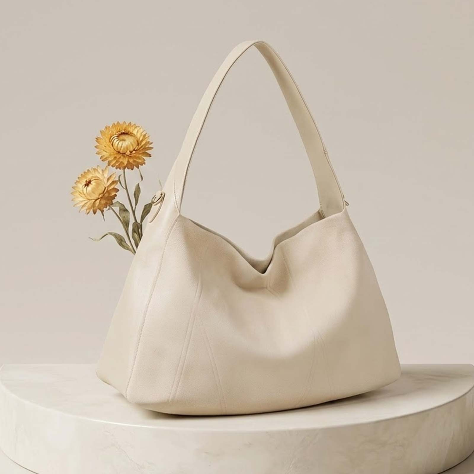 Un bolso hobo de cuero color crema se sienta en un pedestal de mármol junto a flores amarillas.