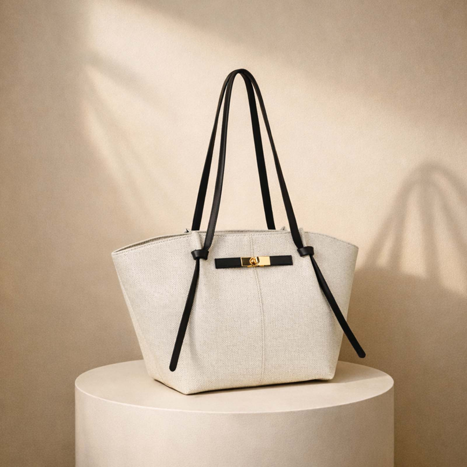 Un bolso tote texturizado color crema con correas negras y herrajes dorados se posa sobre un pedestal.