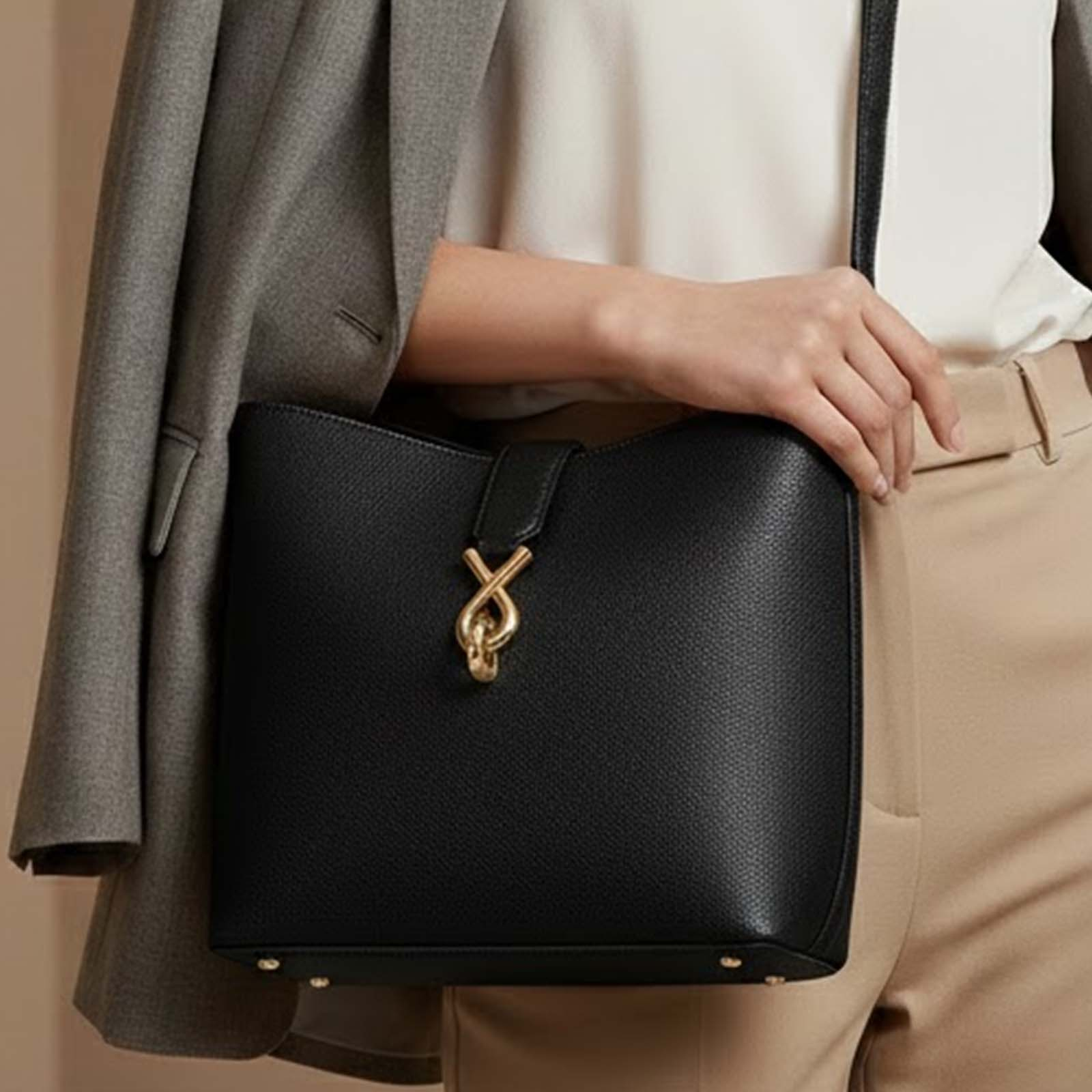 Bolso negro con cierre dorado, llevado por una persona con blazer gris y pantalones beige.