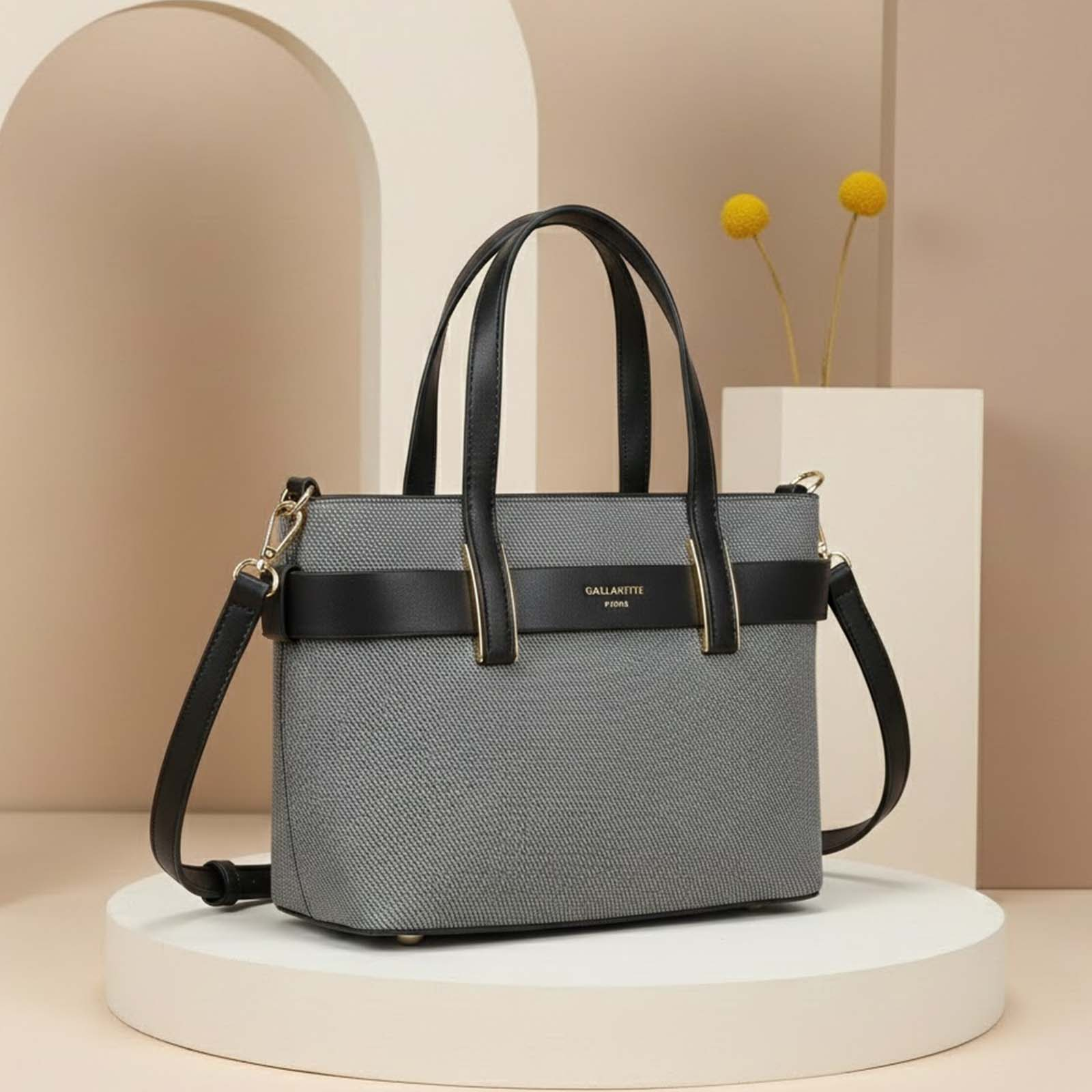 Bolso tote gris con asas y correas negras sobre un pedestal blanco.