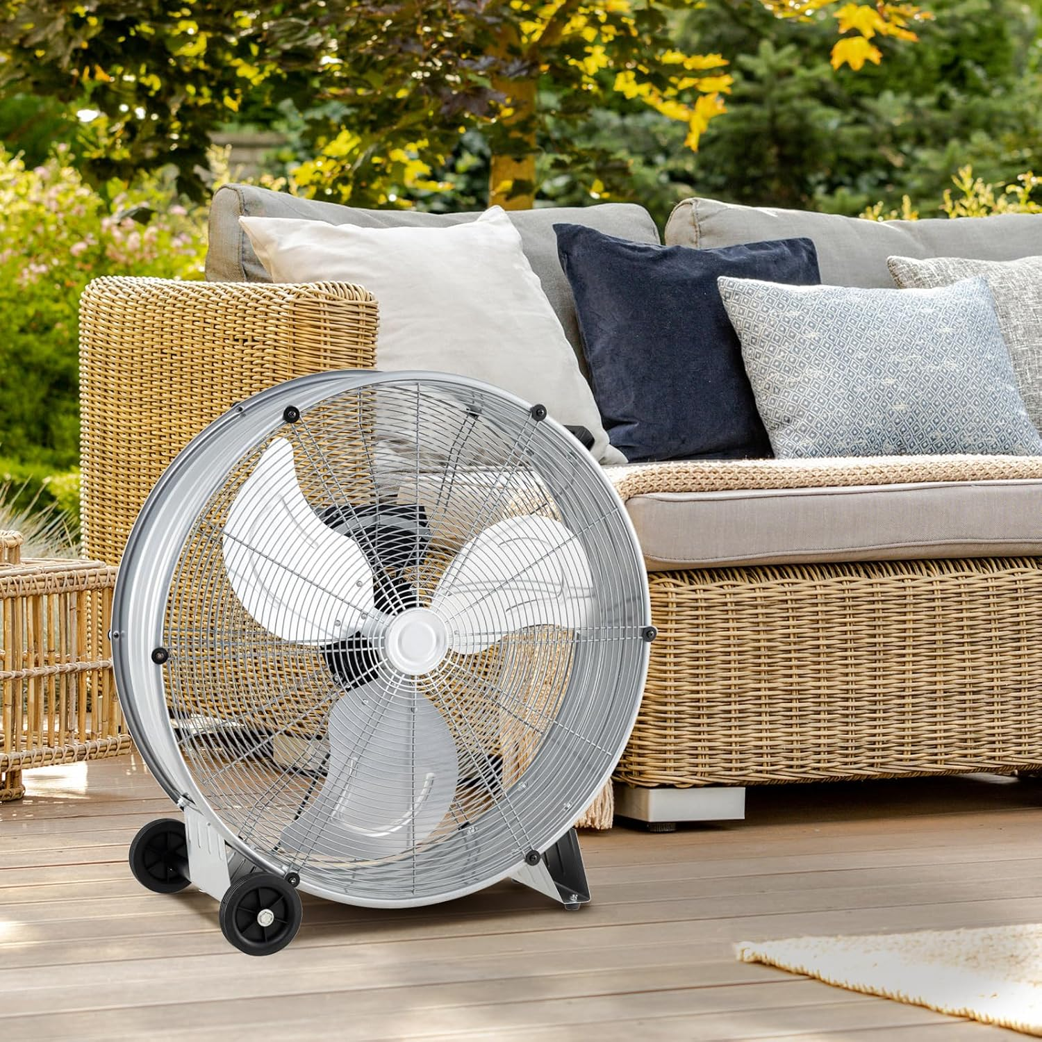 Ein silberner Ventilator steht auf einer Holzterrasse neben einem Rattansofa mit Kissen.