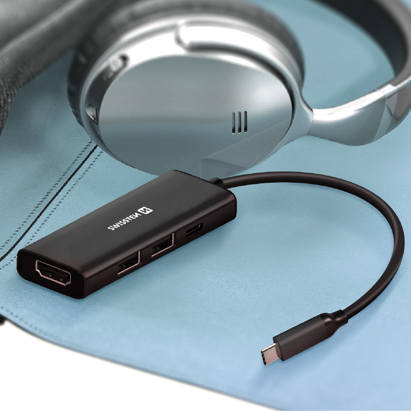 Un hub USB-C nero con porte HDMI e USB è appoggiato su una superficie blu vicino a delle cuffie.
