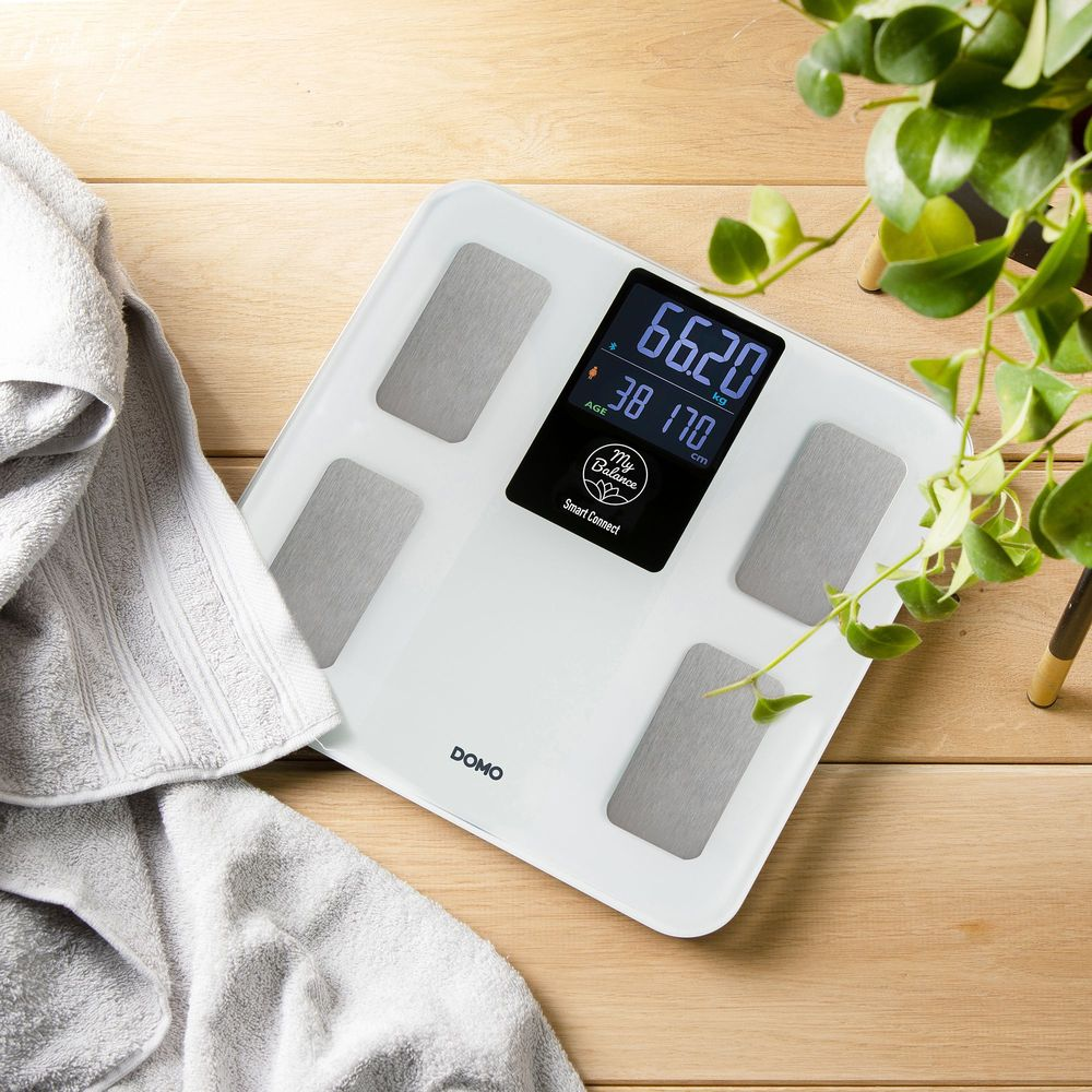 Bilancia digitale bianca mostra 66.20 kg e 38 anni. Asciugamano e pianta nelle vicinanze.