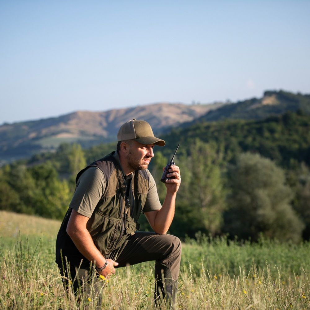 Mann mit Funkgerät kniet in einem Feld mit Hügeln im Hintergrund.