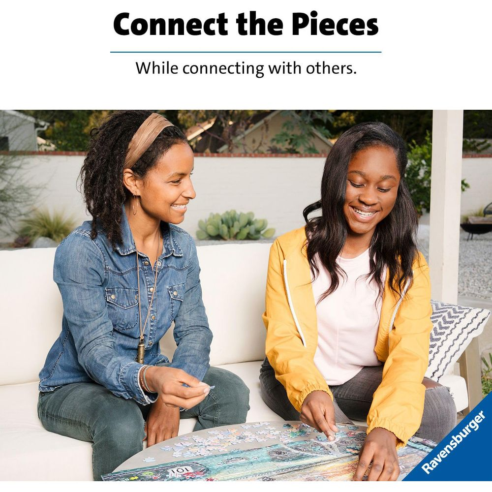 Zwei lächelnde Frauen, eine in Jeans, eine in Gelb, legen draußen auf einer Couch ein Puzzle.