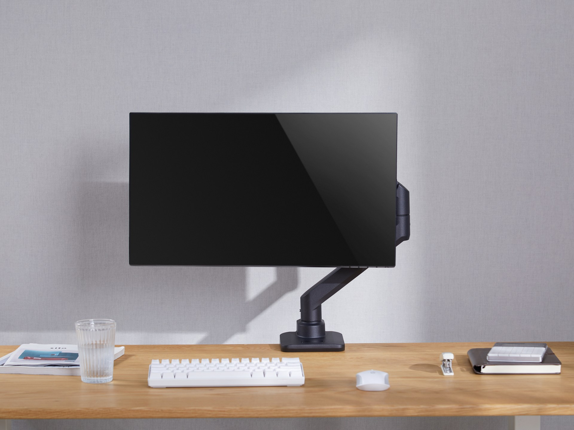 Een zwarte monitor op een bureau met toetsenbord, muis, glas en notitieboek.