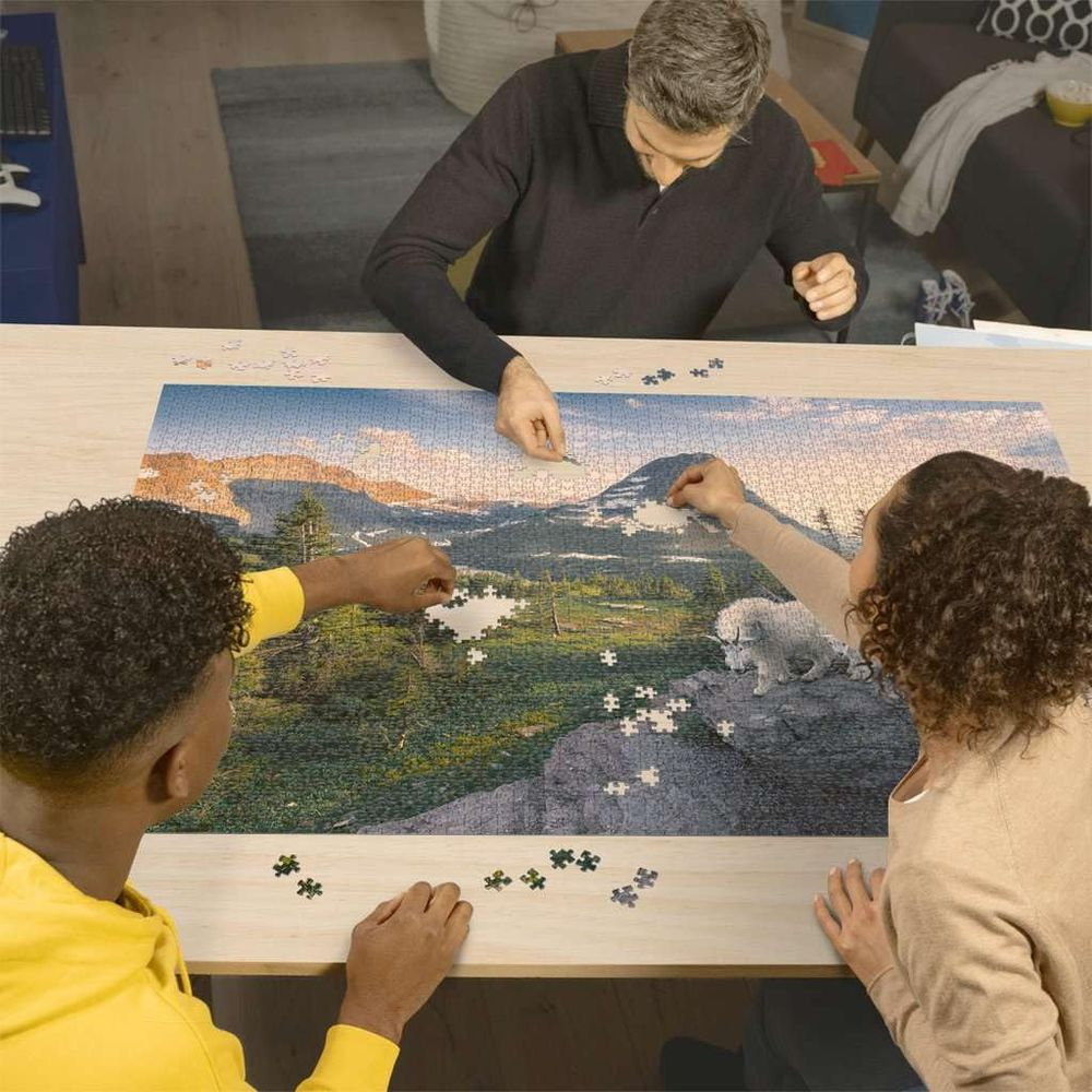 Trois personnes assemblent un grand puzzle de paysage de montagne avec un chien blanc.
