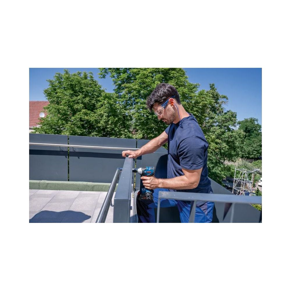 Mann bohrt in einem Geländer auf einem Balkon mit Bäumen und blauer Himmel.