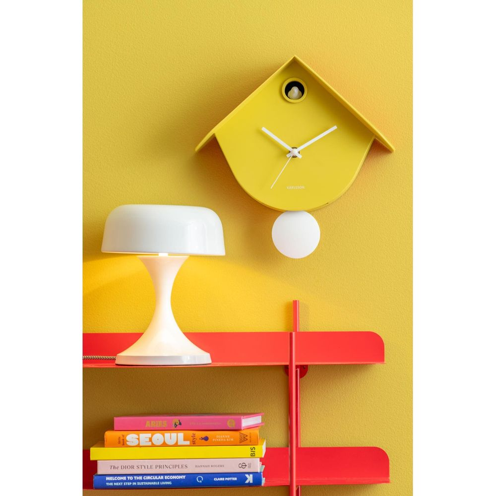 Eine gelbe Vogelhausuhr hängt an einer gelben Wand über einer weißen Lampe und Büchern auf einem roten Regal.