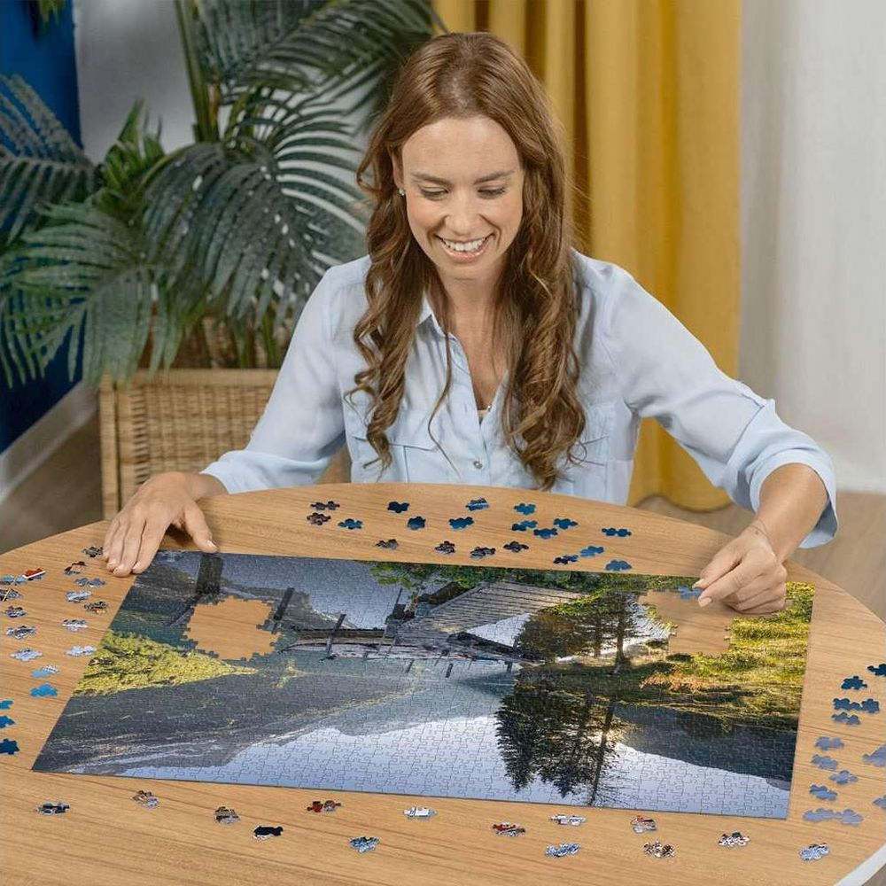 Femme assemblant un puzzle de lac de montagne avec un pont. Beaucoup de pièces sur table.
