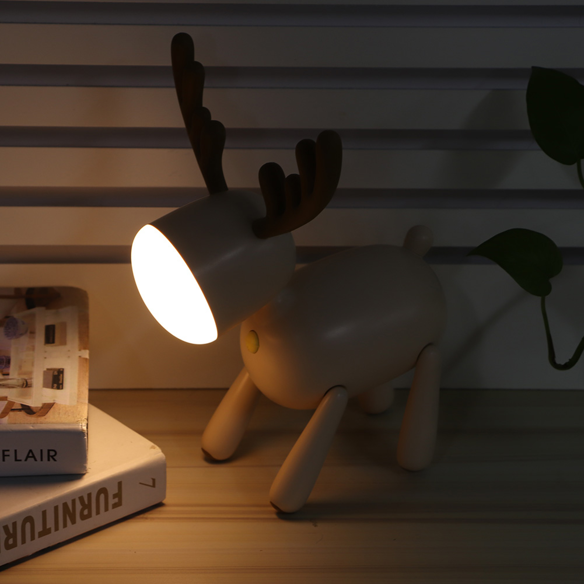 Eine weiße Rentierlampe mit Hörnern steht auf Büchern. Links daneben ist eine Pflanze.