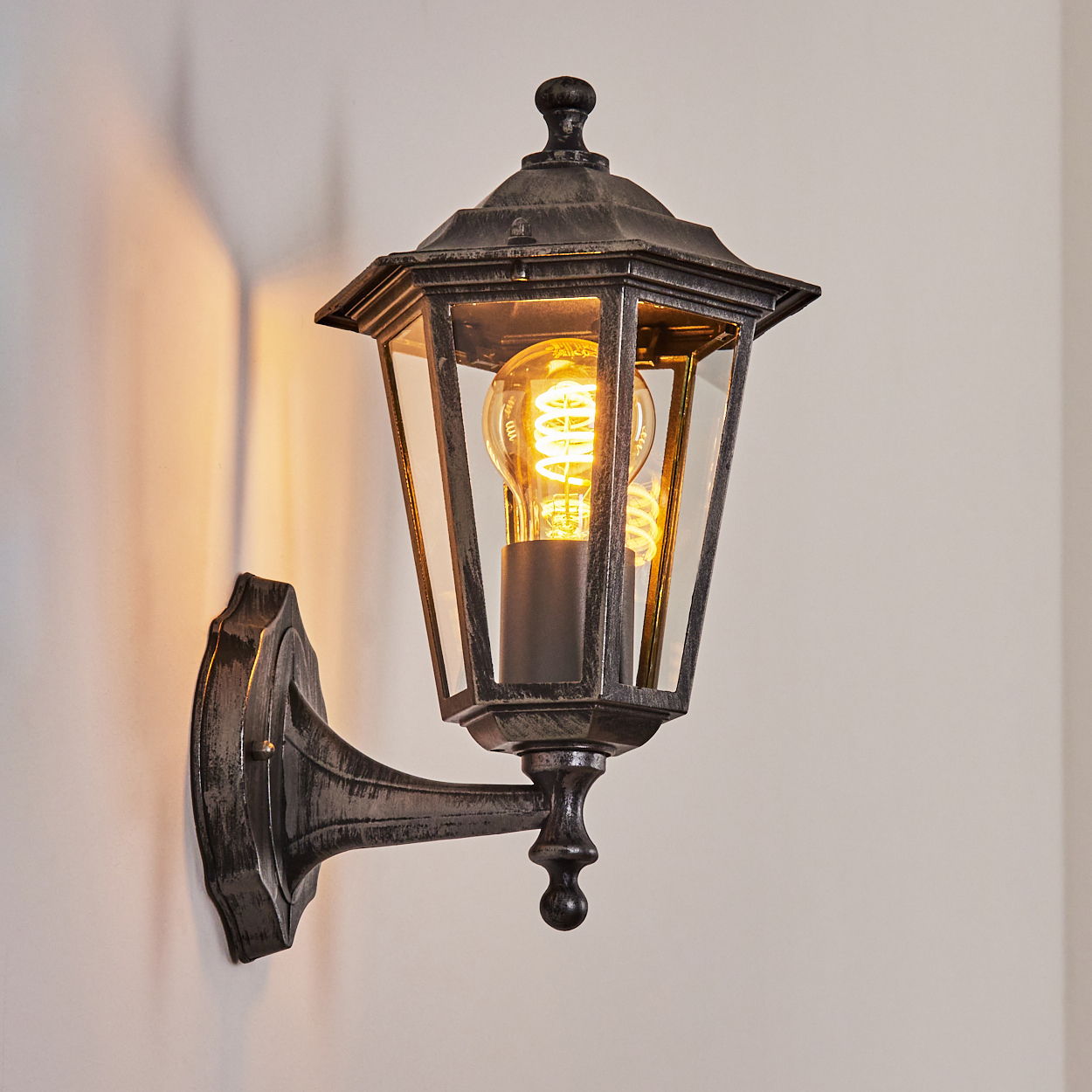 Eine graue Wandleuchte im Laternenstil mit sichtbarem Glühfaden und warmem Licht.