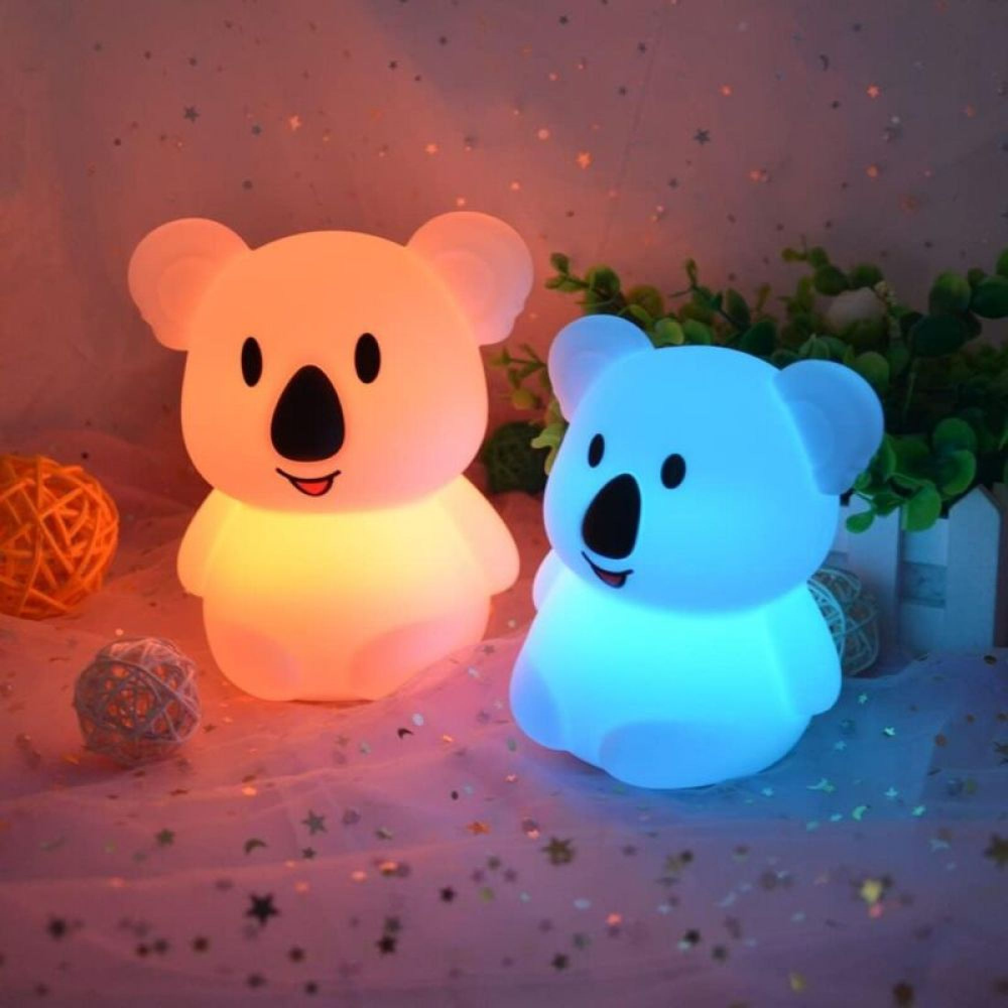 Zwei niedliche Koala-Nachtlichter leuchten in Orange und Blau auf einem Sternenmuster-Tuch.