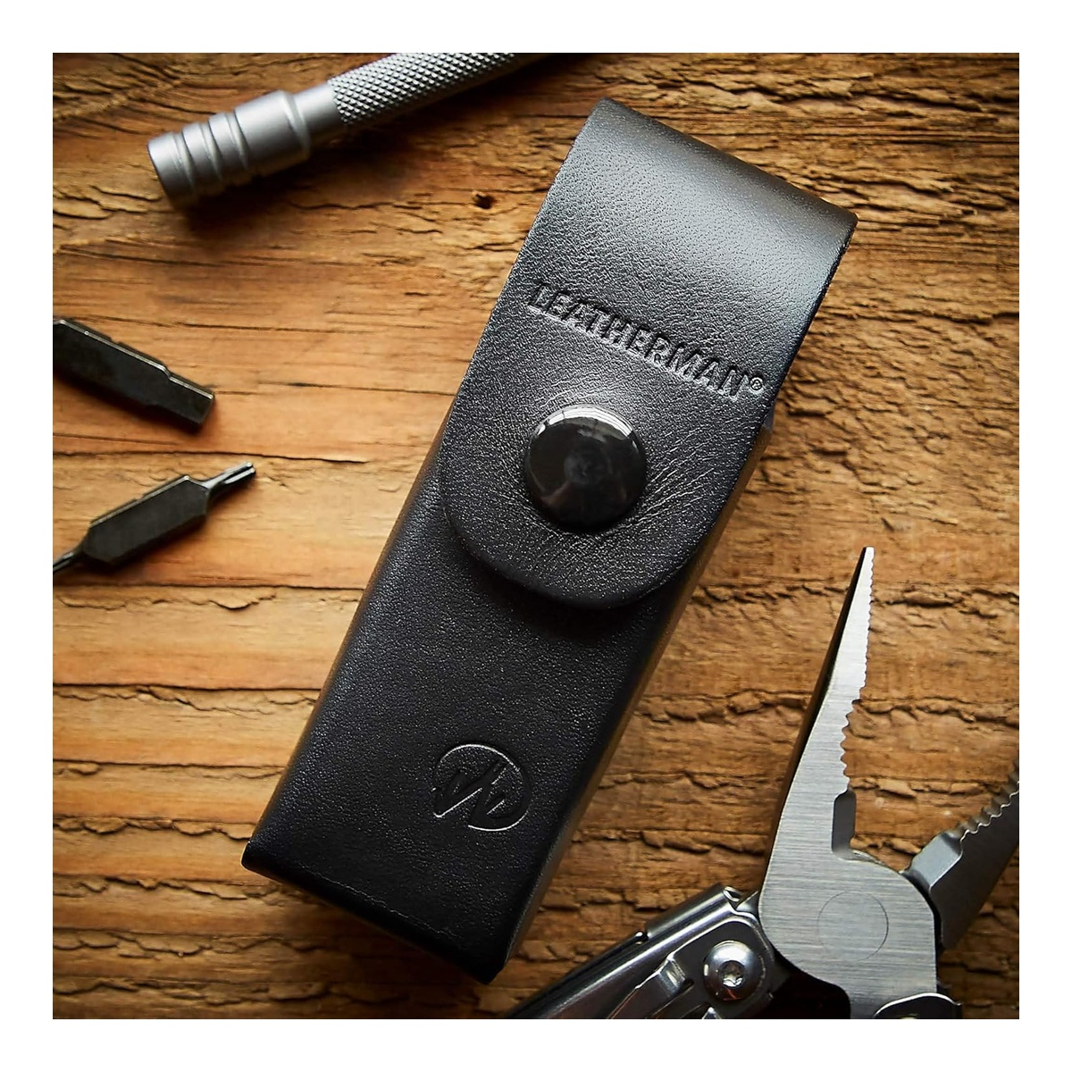 Ein schwarzes Lederetui mit Leatherman-Logo, daneben Zangen und Werkzeugbits auf Holz.