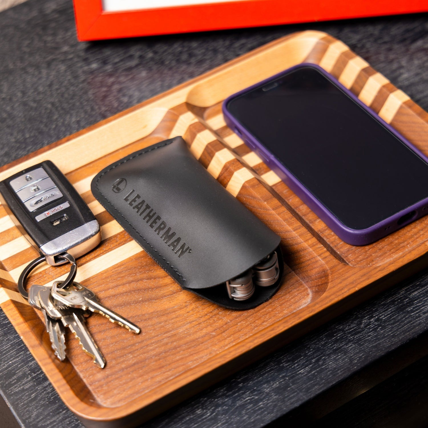Ein Holztablett enthält Schlüssel, ein Leatherman-Werkzeug und eine lila Handyhülle mit Handy.