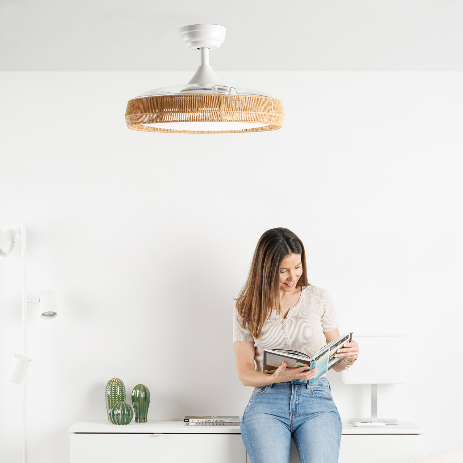 Una mujer con jeans y blusa clara lee un libro, junto a jarrones de cactus.