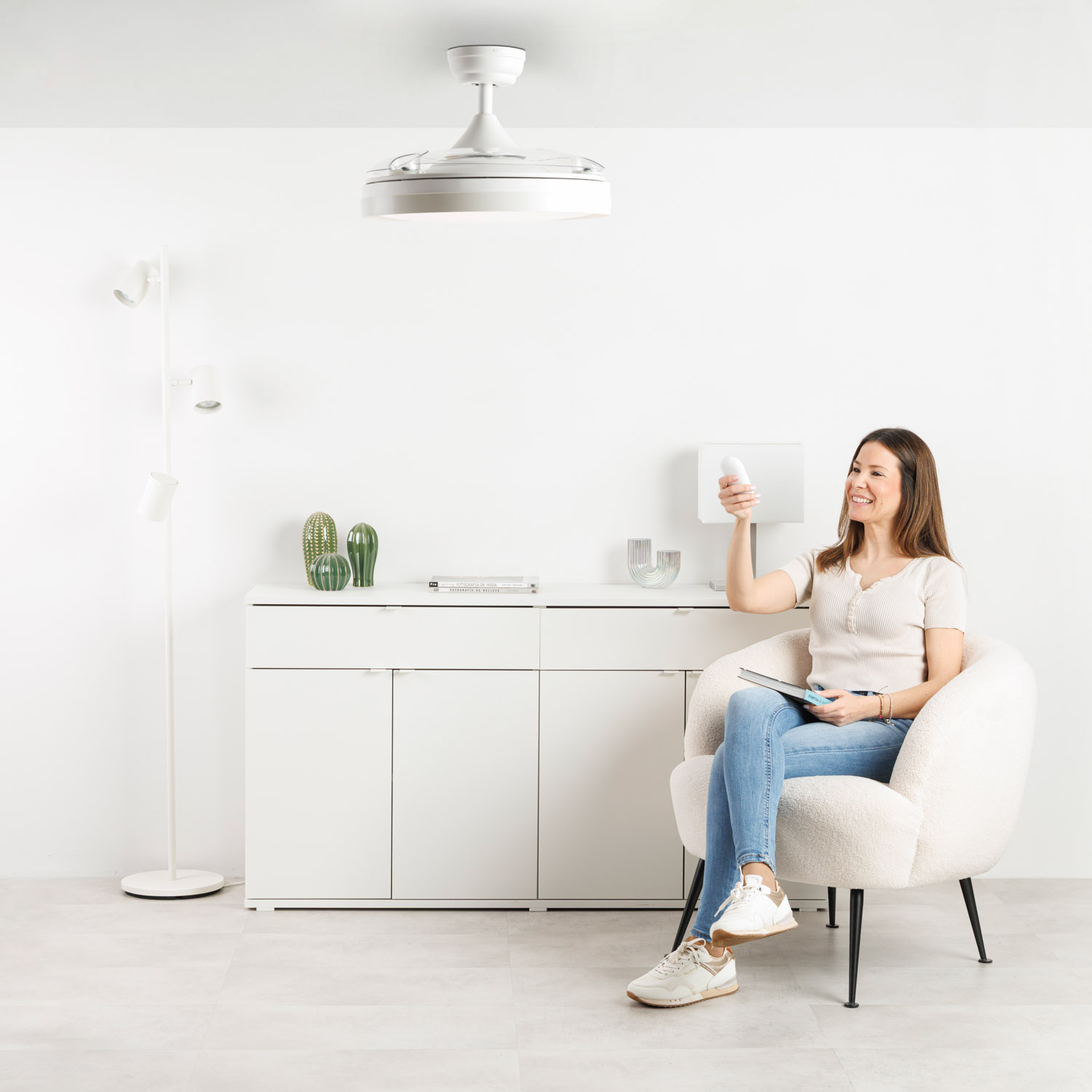 Mujer en sillón sostiene control remoto. Ventilador de techo y mueble atrás.