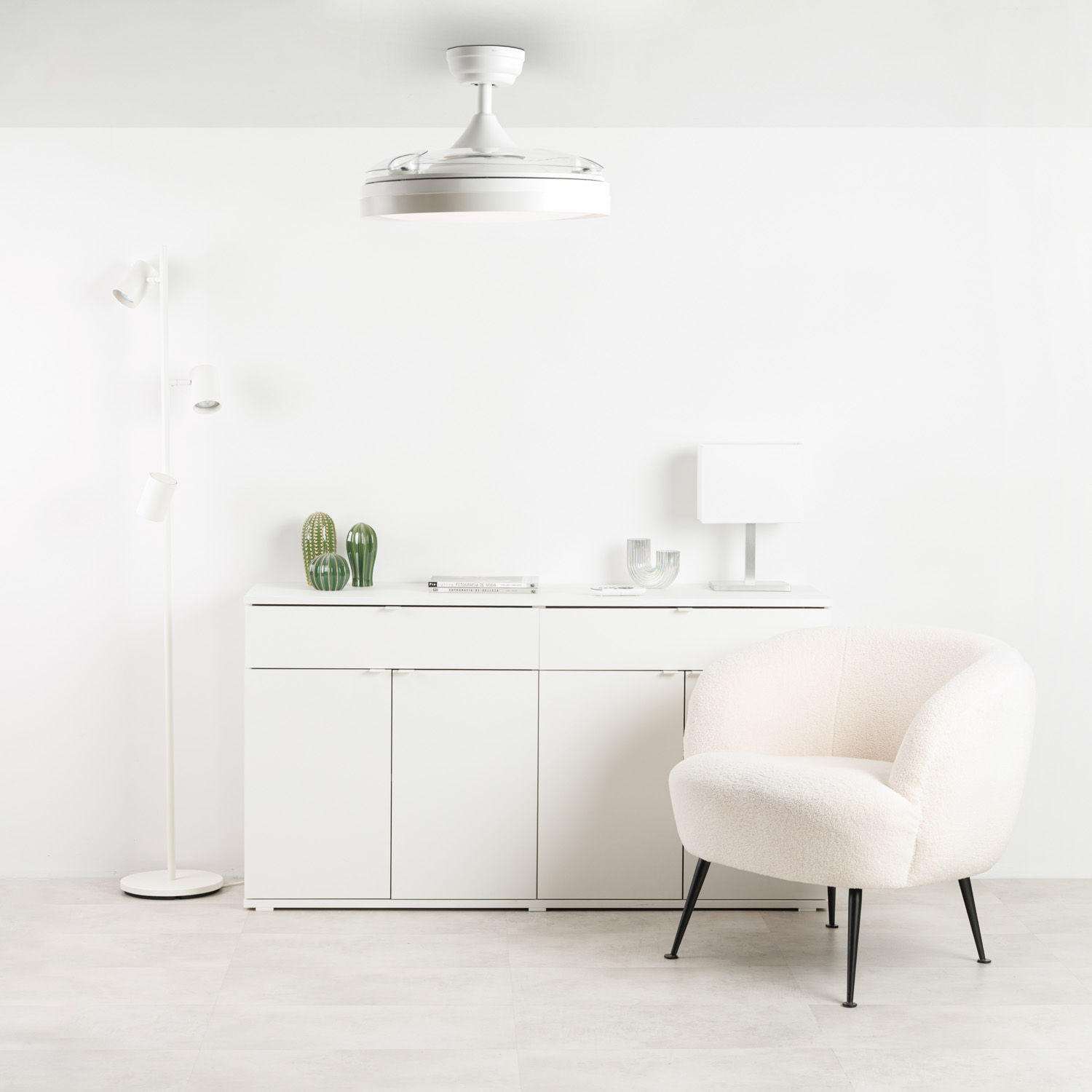 Una habitación blanca con un mueble blanco, sillón de peluche, lámpara y decoración de cactus.