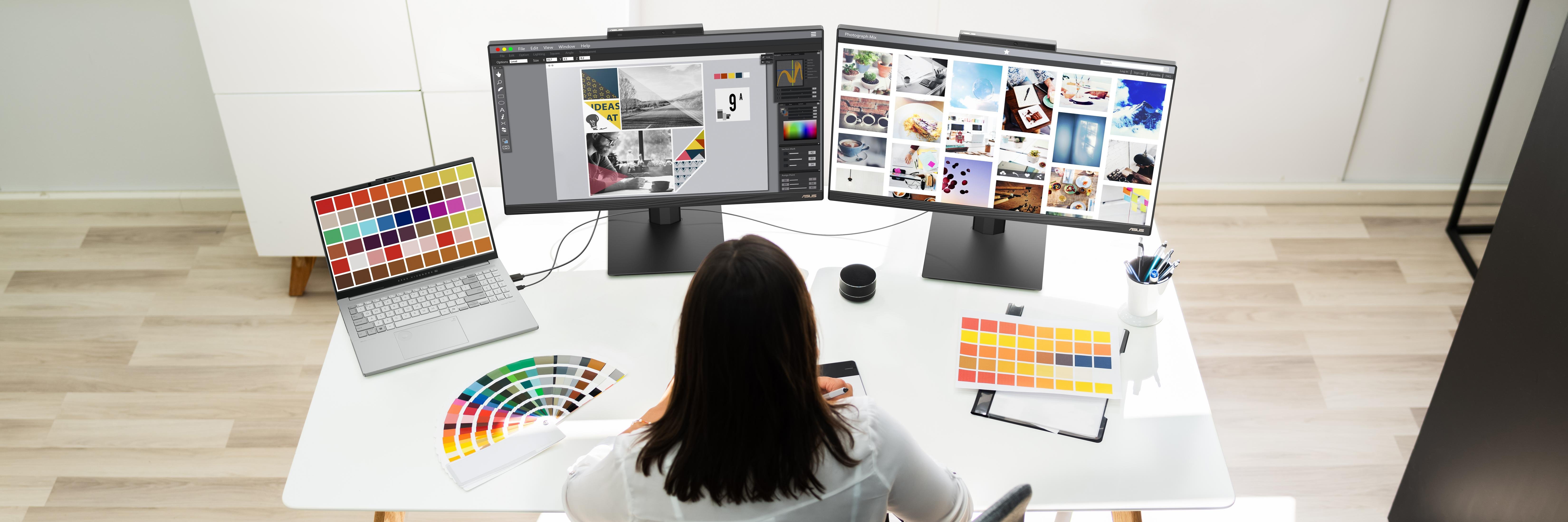 Graphic designer at a desk with dual monitors, laptop, color palettes, and pens.