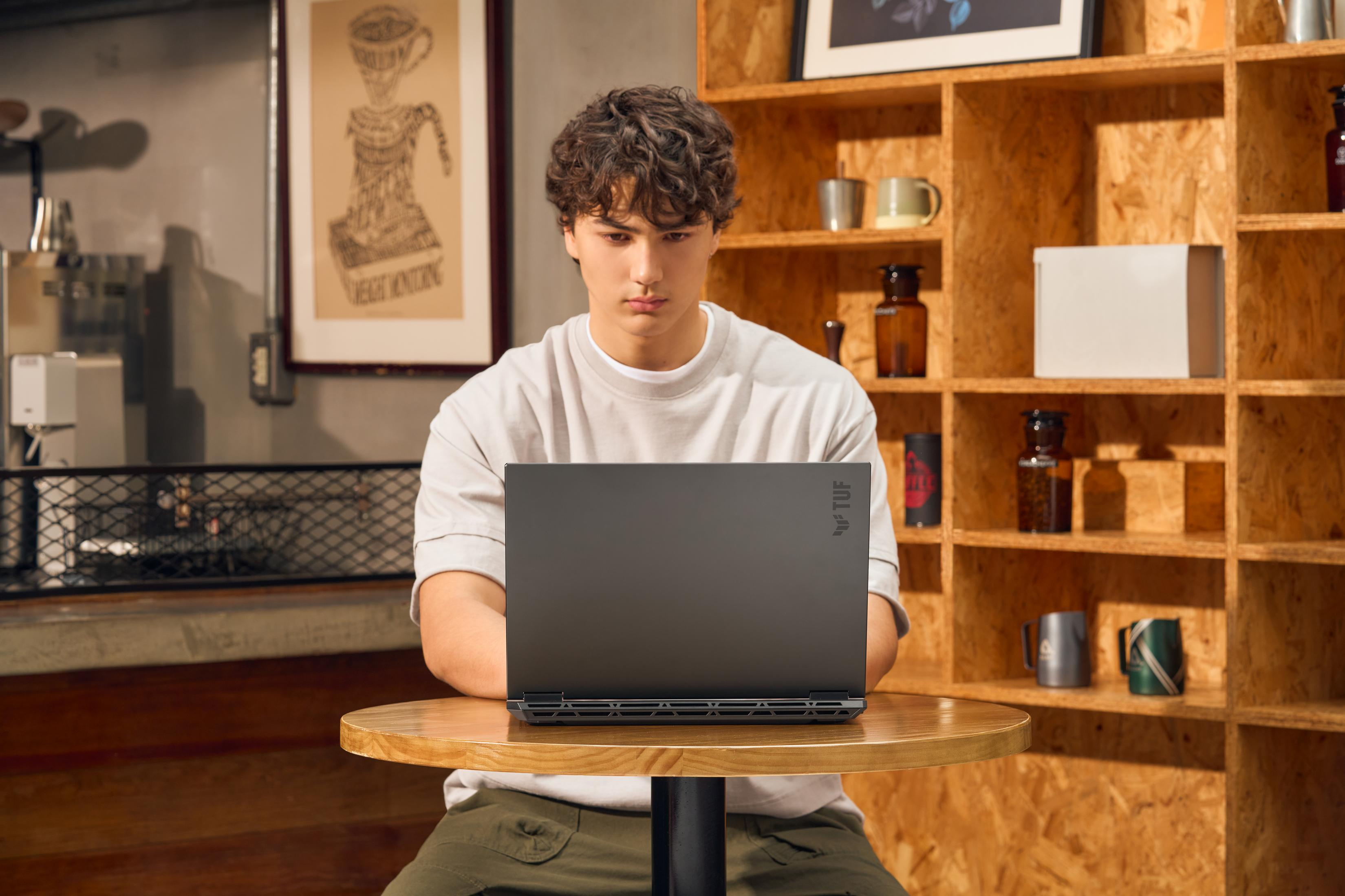 A young man sits at a table using a laptop. Shelves with items are in the background.
